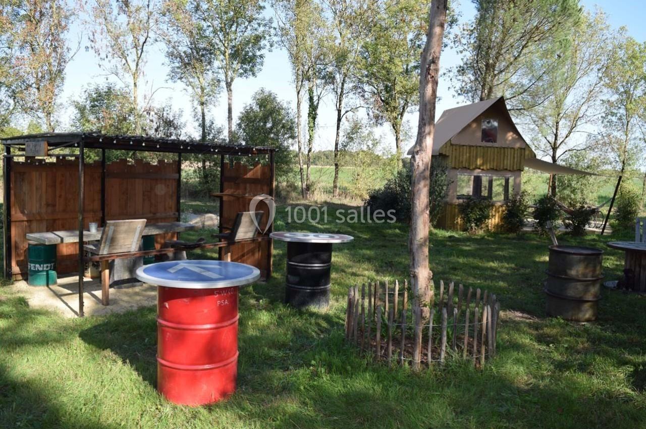 Espace extérieur avec tables fabriquées à partir de tonneaux, abri en bois et petite cabane au fond, entourés d'arbres.