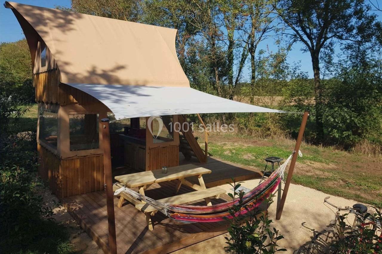 Cabane en bois avec terrasse ombragée, hamac, table de pique-nique et vue sur un environnement naturel verdoyant.