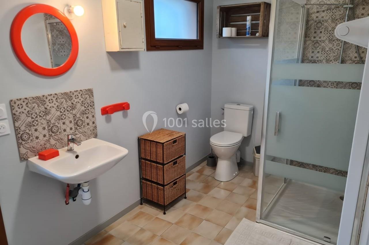 Salle de bain avec lavabo, miroir rond rouge, douche vitrée, toilettes et meubles en osier sur carrelage beige.