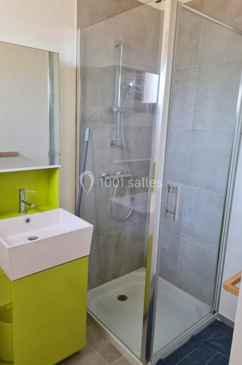 Salle de bain moderne avec douche vitrée, lavabo sur meuble vert et miroir, sol en carrelage clair.