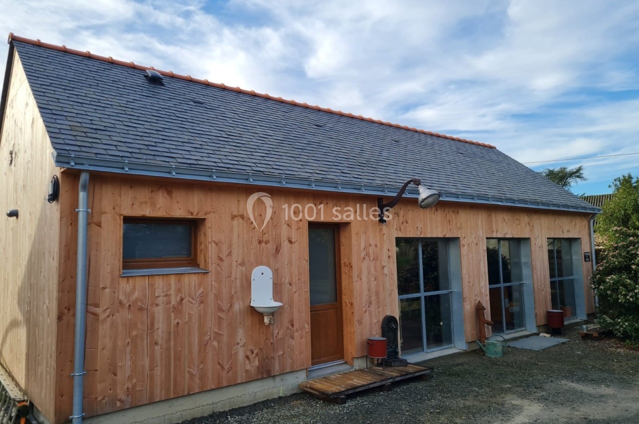 Maison en bois avec toit en ardoise, grandes baies vitrées et petite cour gravillonnée sous un ciel partiellement nuageux.