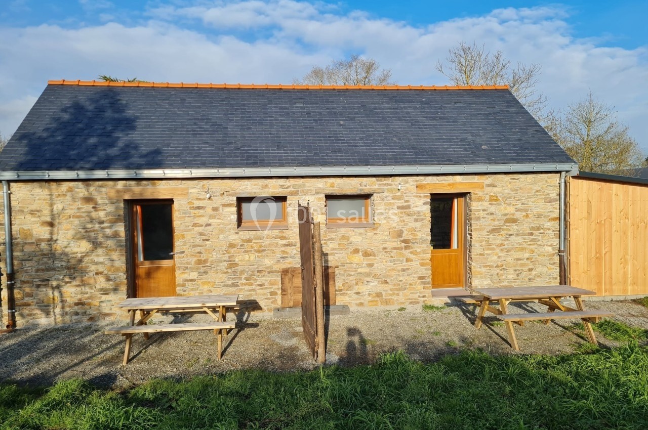 Petite maison en pierre avec toit en ardoise, deux portes en bois, tables de pique-nique et ciel dégagé en arrière-plan.