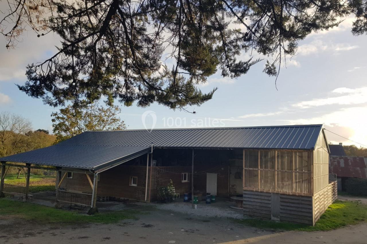 Bâtiment agricole en bois et métal avec un toit en pente, entouré d'arbres et baigné par une lumière naturelle.