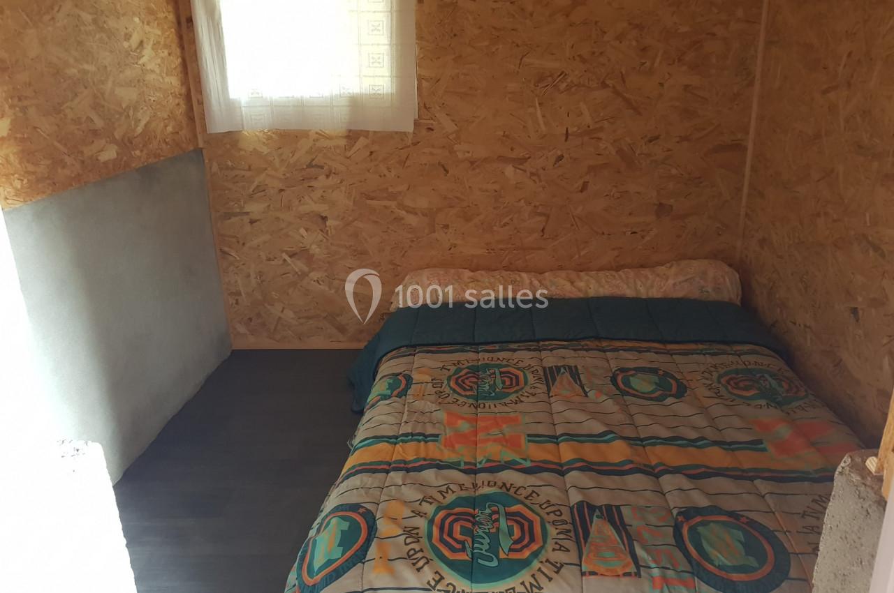 Lit double avec une couverture colorée dans une petite chambre aux murs en panneaux de bois, éclairée par une fenêtre.