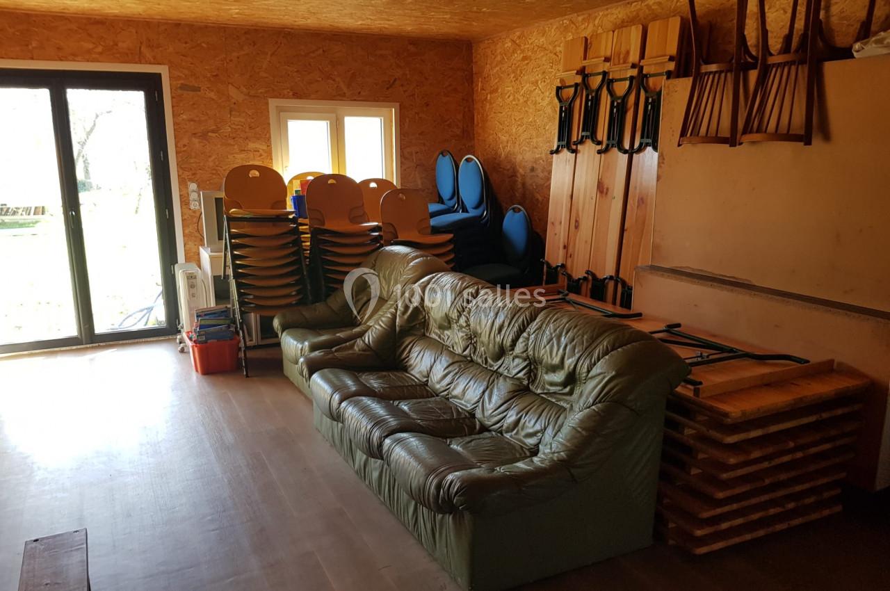 Salle avec canapé en cuir vert, chaises empilées, tables pliées et grande baie vitrée donnant sur l'extérieur.