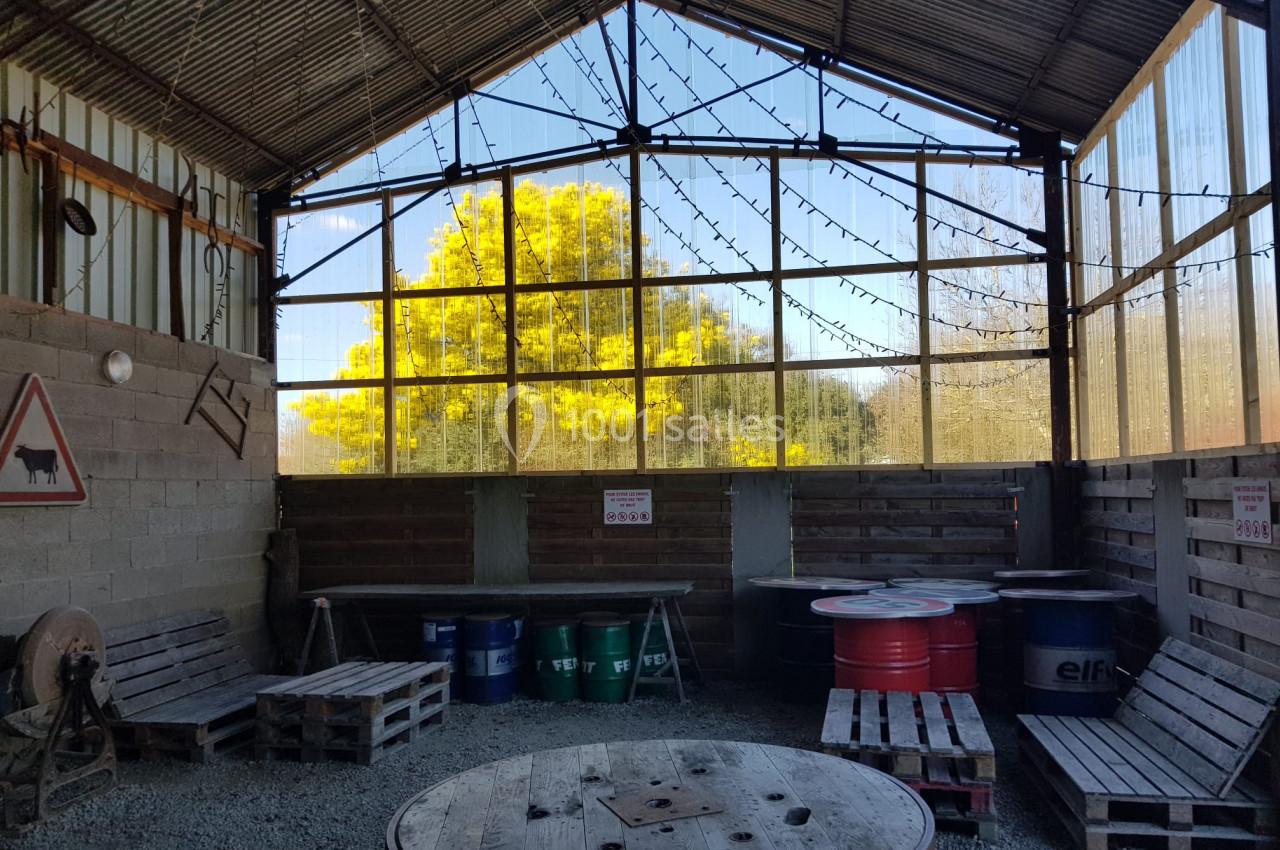 Intérieur d'un hangar avec des palettes en bois, des tonneaux colorés et une grande baie vitrée donnant sur un arbre jaune.