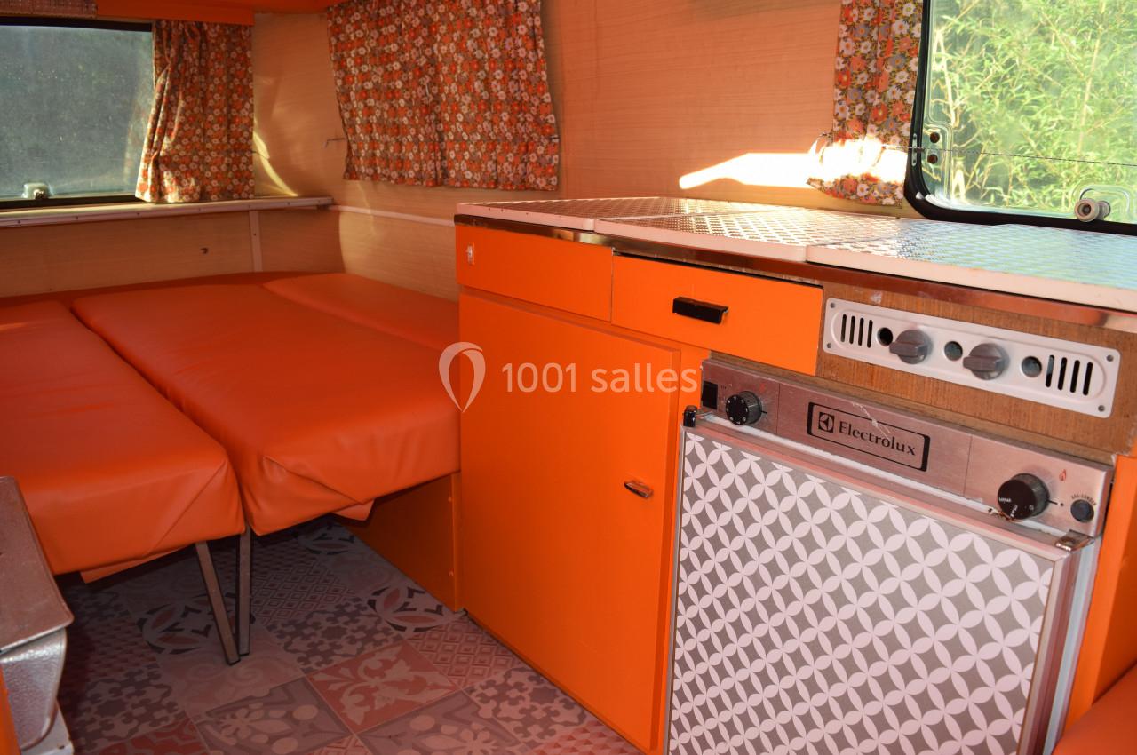 Intérieur d'une caravane rétro avec banquettes orange, rideaux à motifs et kitchenette équipée d'un réfrigérateur.