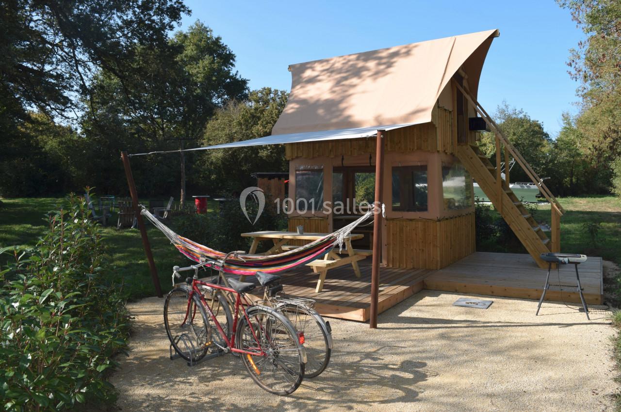 Cabane en bois avec terrasse, hamac, table de pique-nique et vélos, située dans un cadre naturel verdoyant.