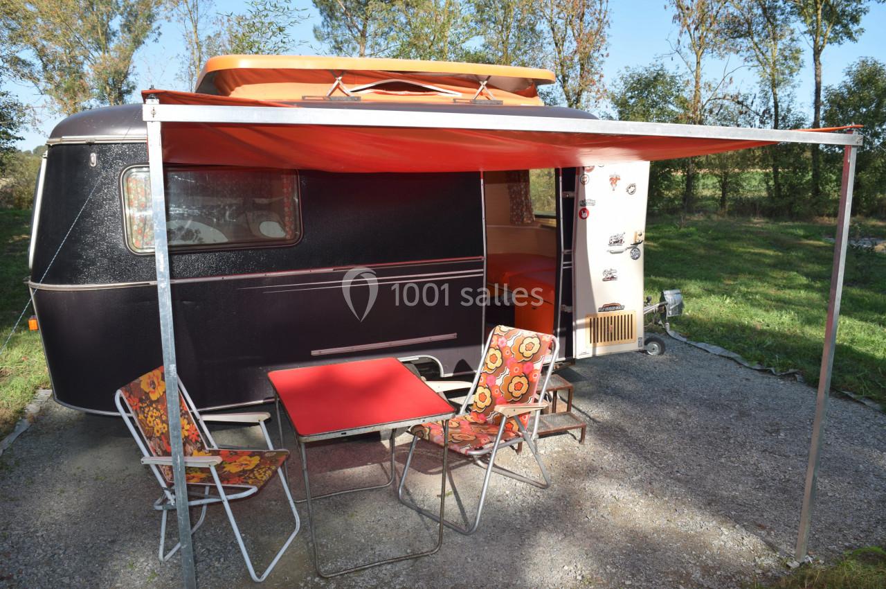 Caravane noire avec auvent orange, table et chaises installées sur un emplacement ensoleillé entouré de verdure.