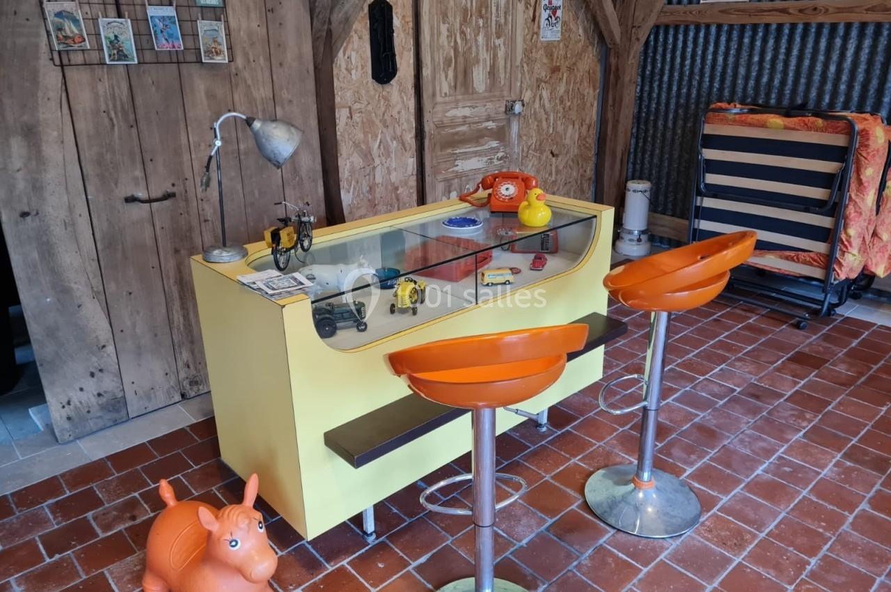 Comptoir jaune avec objets rétro, deux tabourets orange, sol en carreaux rouges et décor rustique en bois et métal.
