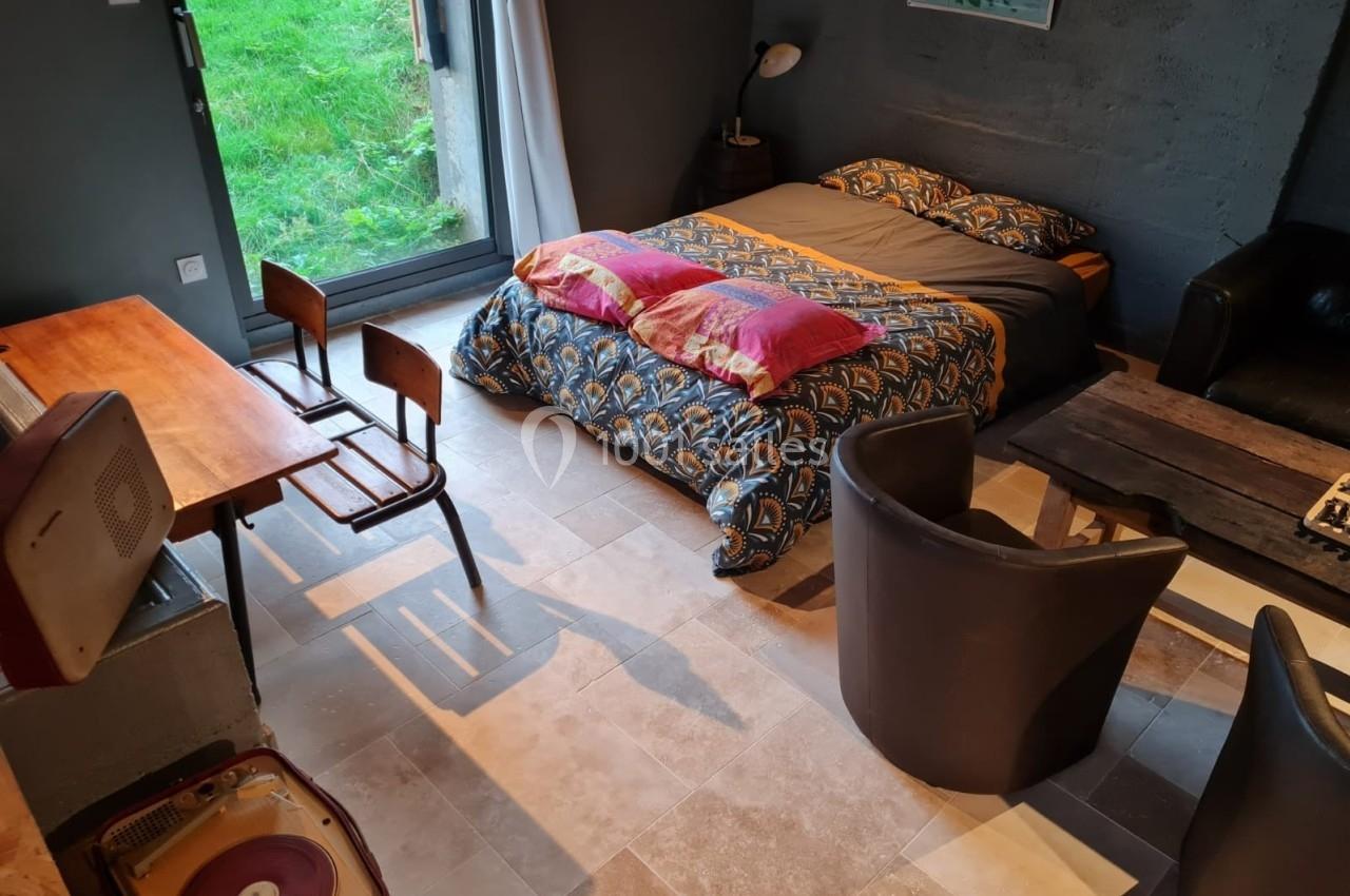 Chambre avec lit double, table en bois, chaises, fauteuils, tourne-disque et grande fenêtre donnant sur un jardin.