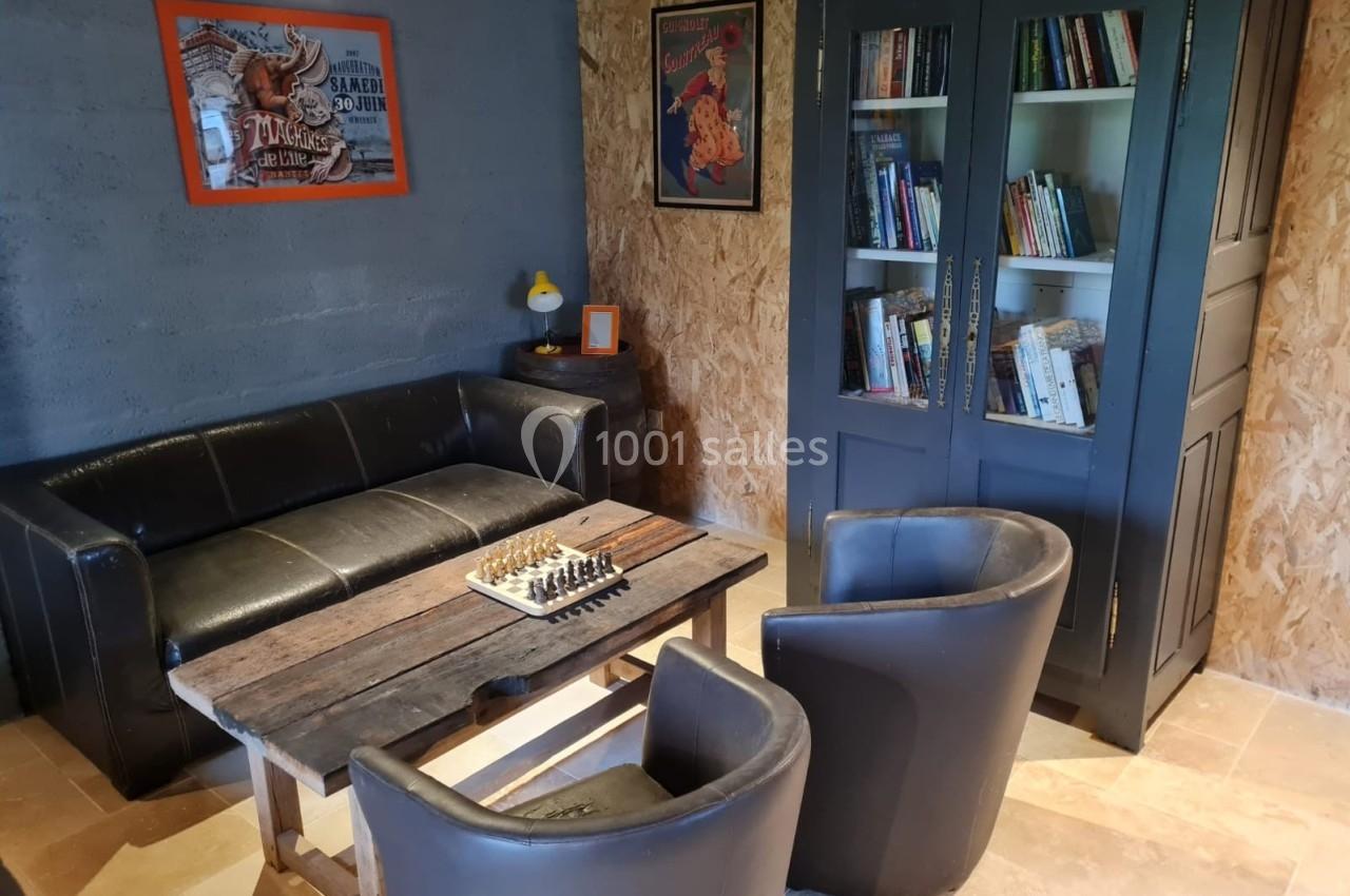 Espace de détente avec canapé, fauteuils, table basse avec échiquier, bibliothèque et décorations murales.