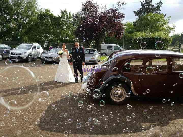 Un couple en tenue de mariage marche près d'une voiture ancienne, entouré de bulles, dans un cadre extérieur verdoyant.