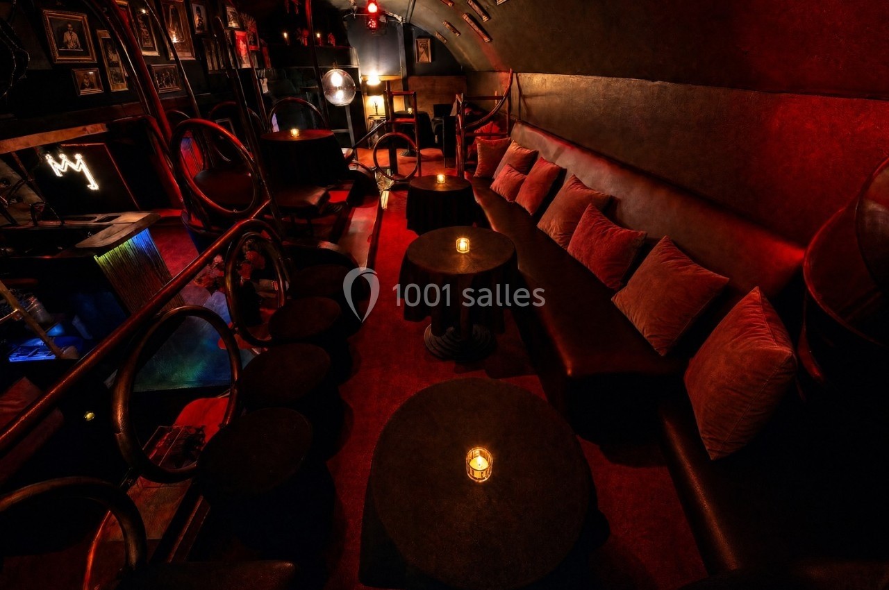 Salle de bar tamisée avec banquettes, coussins, petites tables rondes et éclairage rouge chaleureux.