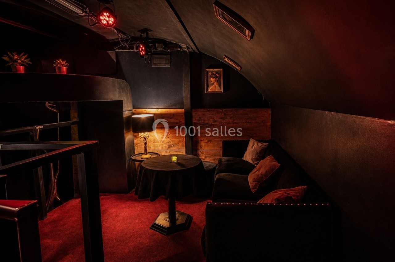 Espace lounge sombre avec canapé, table basse, lampe allumée et décoration murale dans une ambiance tamisée.