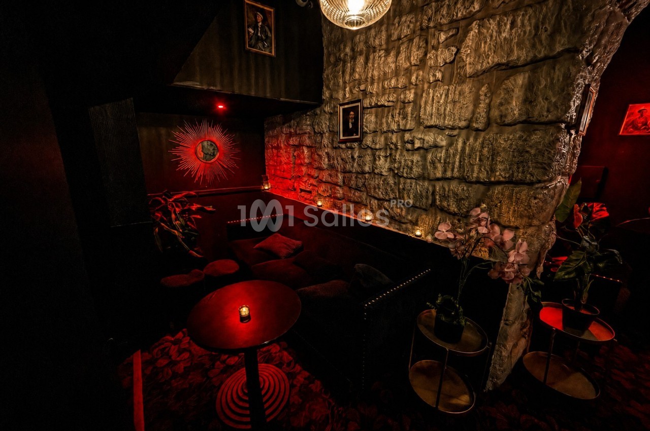 Coin salon sombre avec canapé noir, murs en pierre, éclairage rouge tamisé et petite table avec bougie.