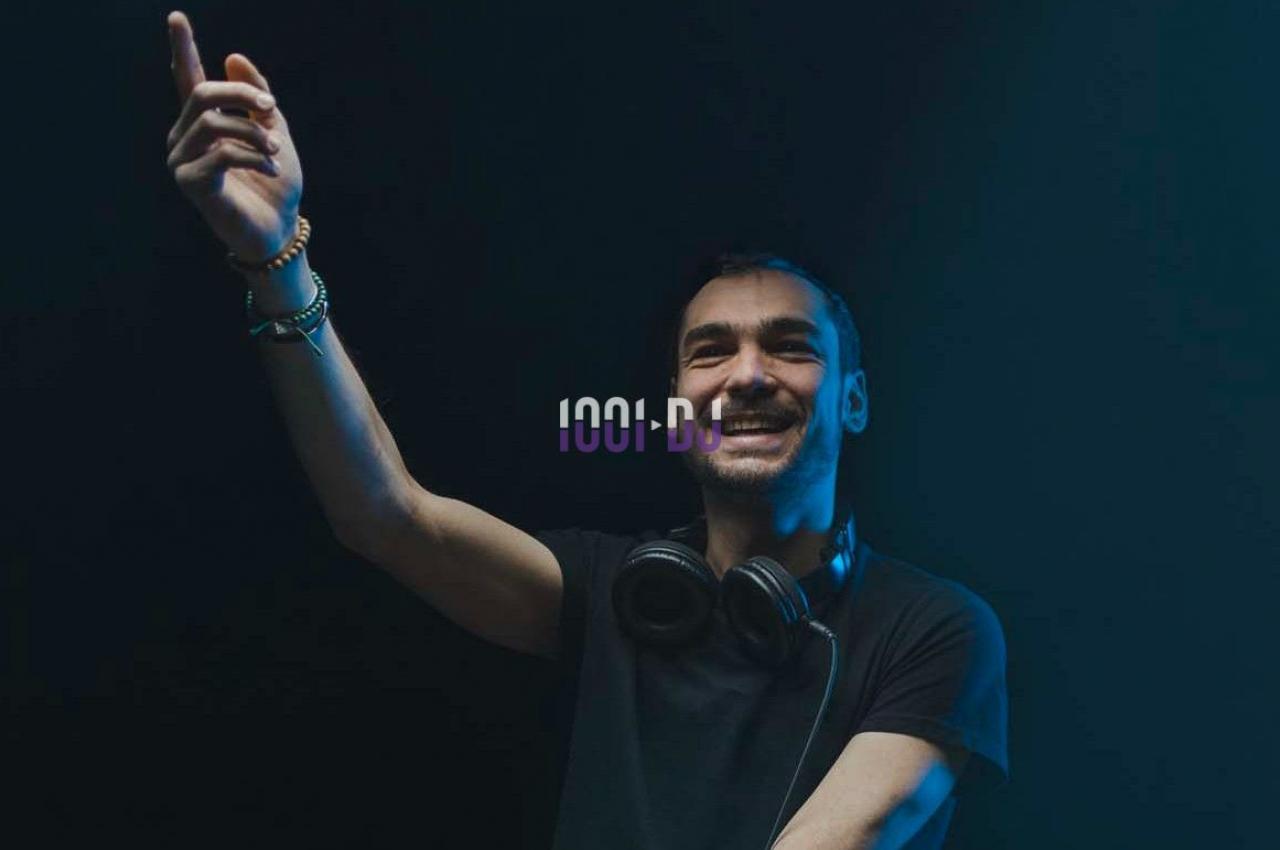Un DJ souriant portant un casque autour du cou lève la main dans une ambiance sombre et lumineuse.