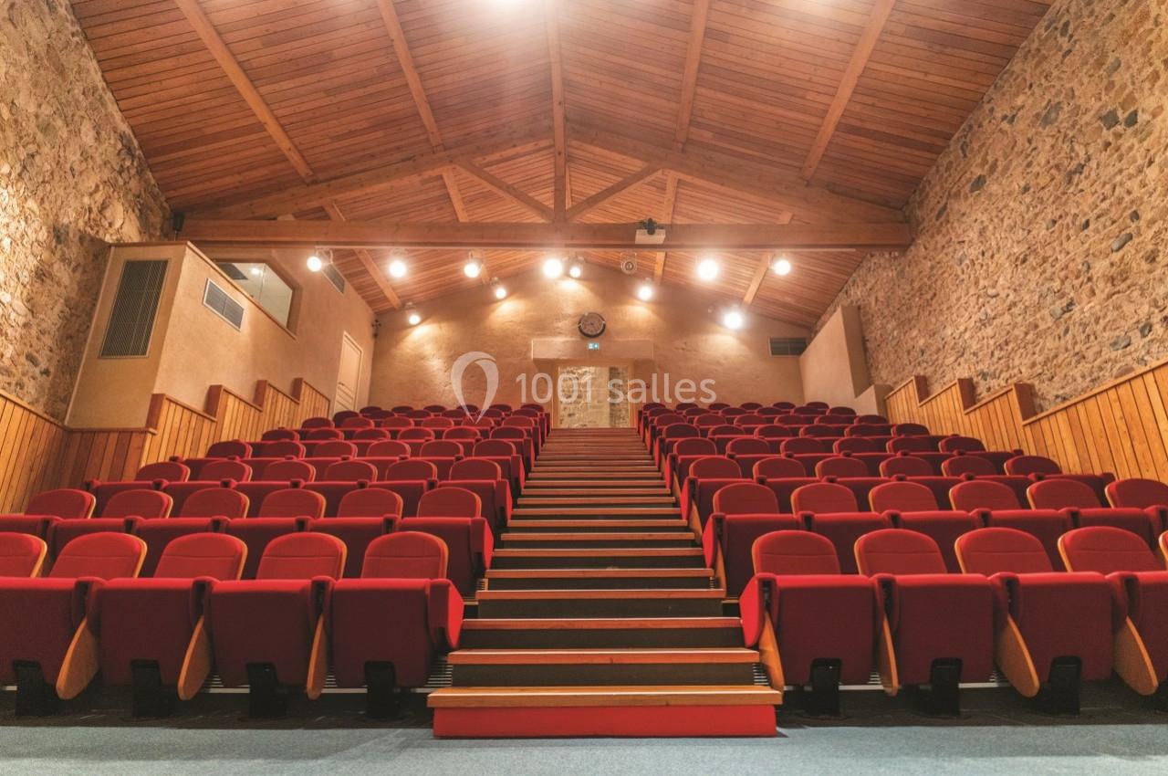 Salle de spectacle avec des rangées de sièges rouges, murs en pierre et plafond en bois éclairé par des spots.