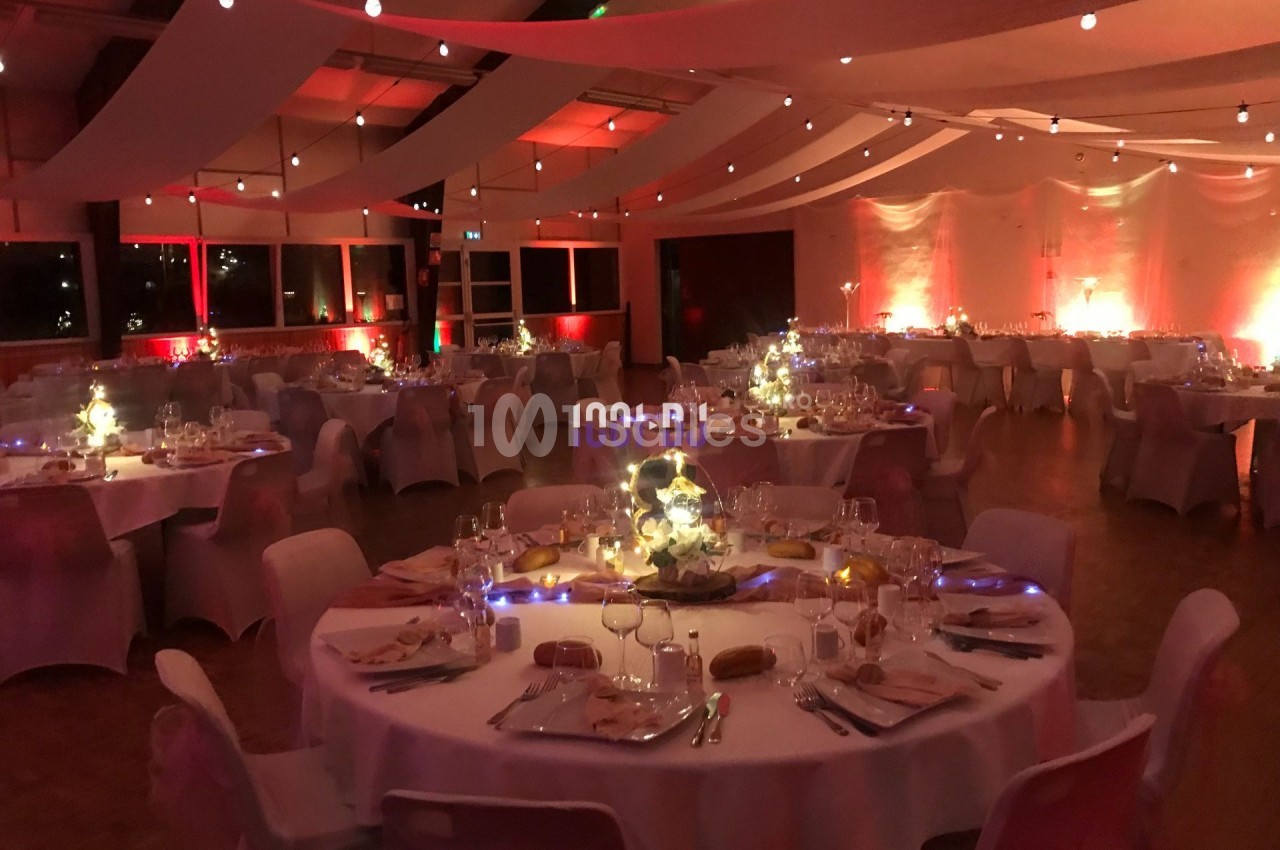 Salle de réception décorée avec des tables rondes dressées, éclairage tamisé et guirlandes lumineuses suspendues.