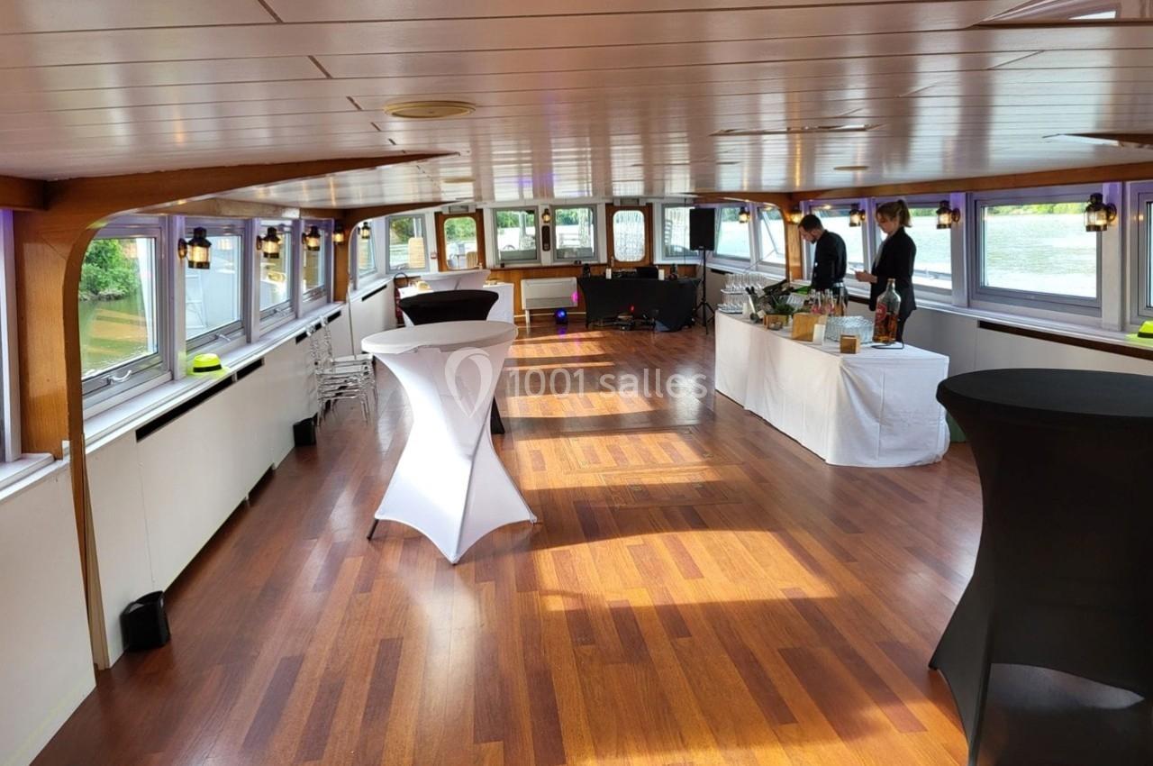 Salle lumineuse sur un bateau avec parquet, tables hautes et buffet installé, prête pour un événement.