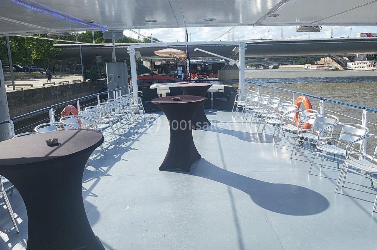 Pont supérieur d'un bateau aménagé avec des tables hautes et des chaises, amarré près d'une rive sous un ciel dégagé.
