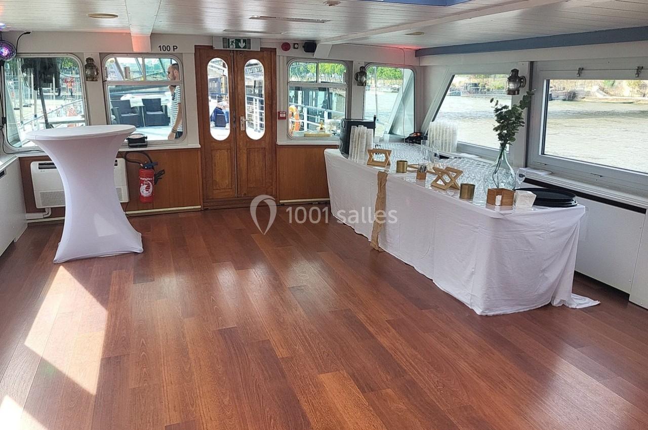 Intérieur d'une salle sur un bateau avec parquet, table drapée pour buffet et vue sur l'extérieur par les fenêtres.