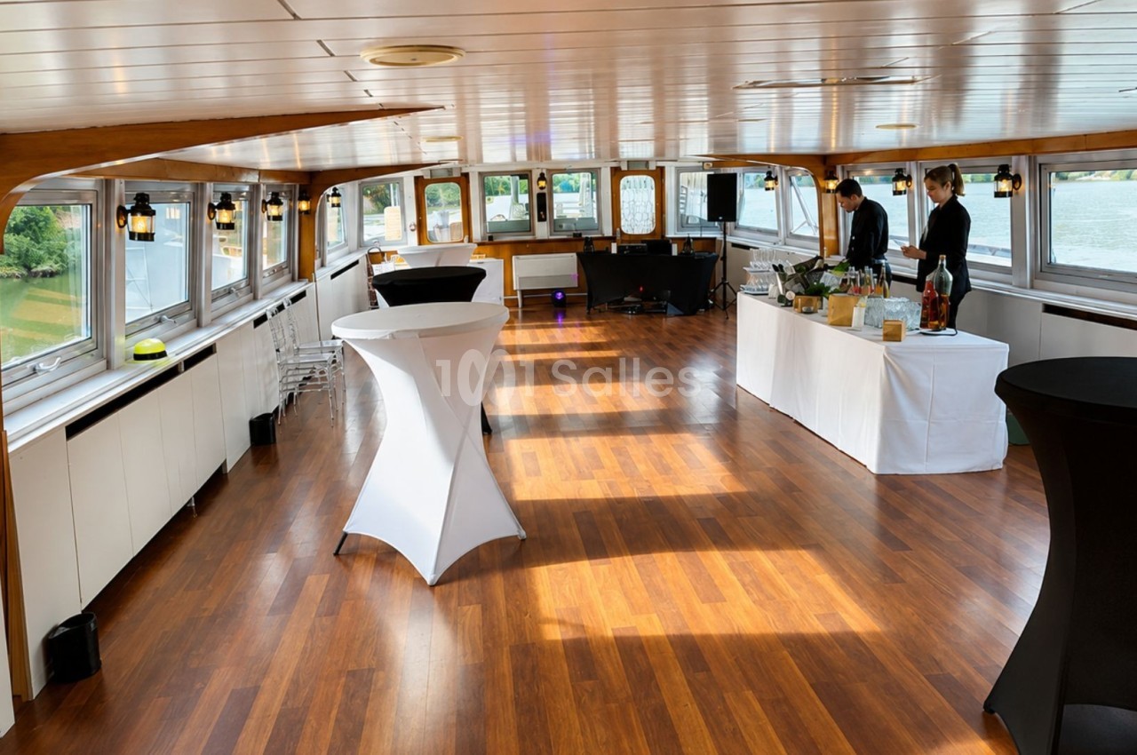 Salle lumineuse avec parquet en bois, tables hautes et buffet préparé, située sur un bateau avec vue sur l'extérieur.