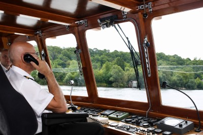 Miniature Location salle Paris 16 (Paris) - Bateau Le Rêve sur Seine #13 Un capitaine en uniforme utilise une radio dans la cabine d'un bateau, avec vue sur une rivière bordée d'arbres.