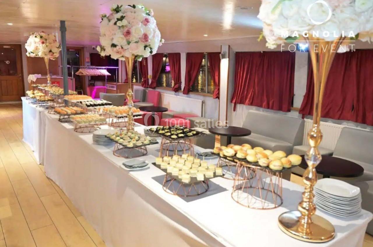 Buffet élégant avec desserts variés, fleurs blanches et roses en décoration, dans une salle lumineuse avec rideaux rouges.