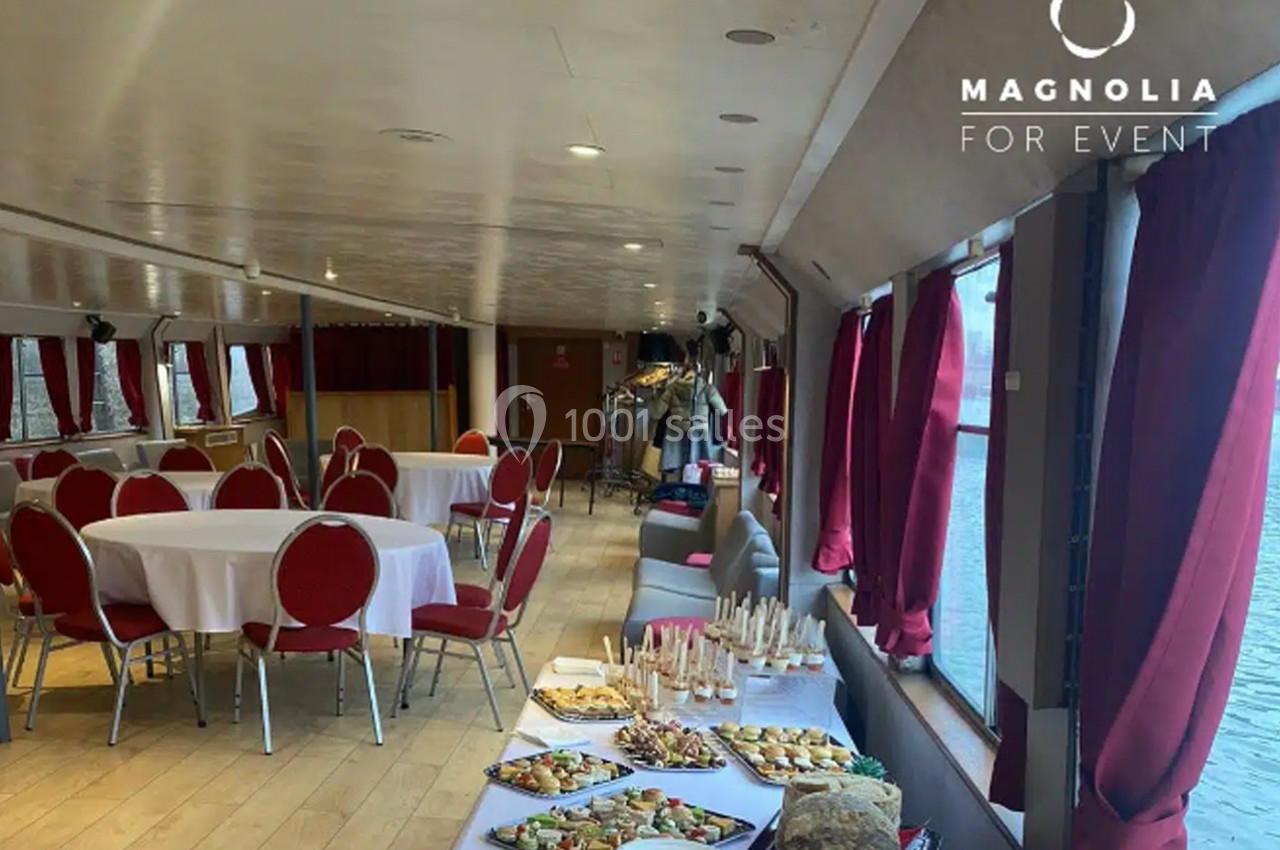 Salle de réception sur un bateau avec tables dressées, chaises rouges et buffet disposé près des fenêtres.