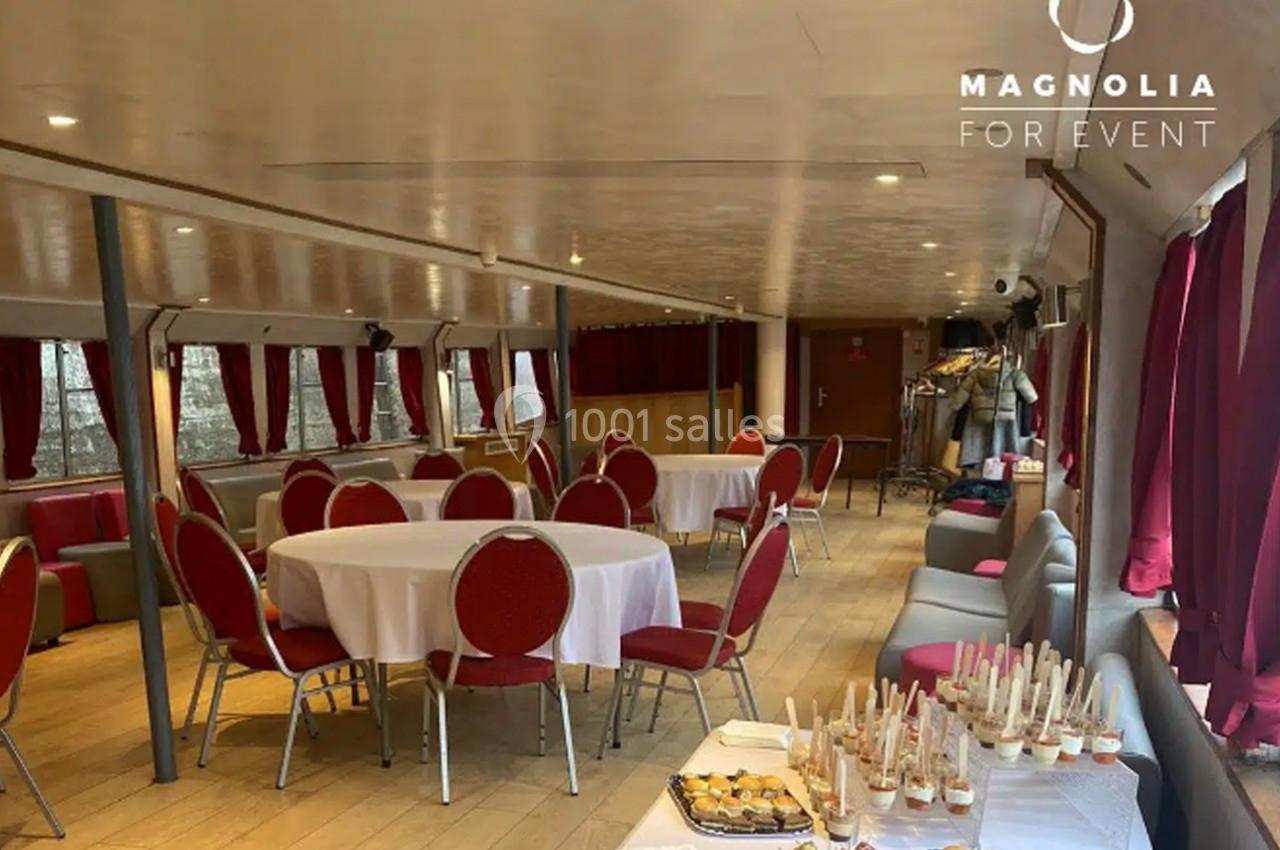 Salle de réception sur une péniche avec tables rondes dressées, rideaux rouges et buffet de desserts au premier plan.