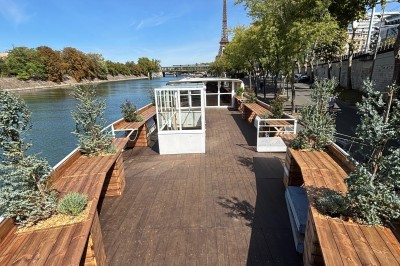 Intérieur d'un bateau avec éclairage vert, tables et chaises, vue sur une terrasse extérieure et des passants.