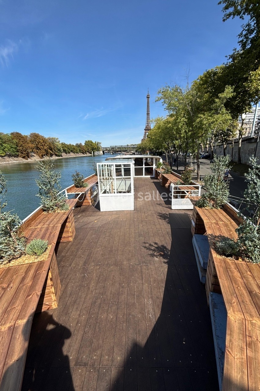 Terrasse en bois aménagée sur une péniche, bordée de plantes, avec vue sur la Seine et la tour Eiffel en arrière-plan.