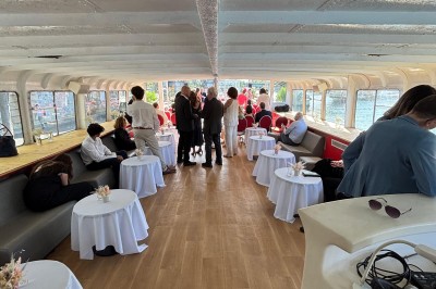 Intérieur d'un bateau avec éclairage vert, tables et chaises, vue sur une terrasse extérieure et des passants.