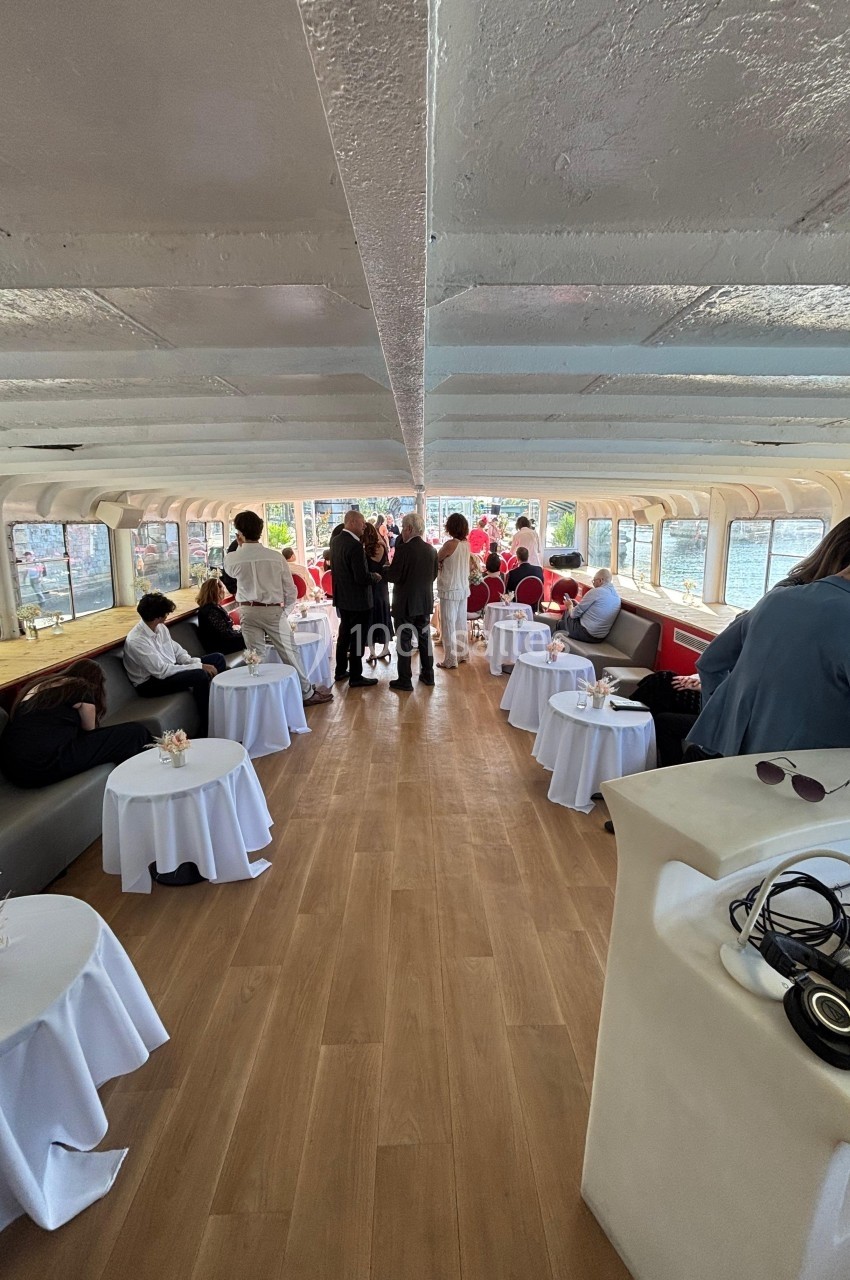 Intérieur d'un bateau avec des tables dressées, des invités debout et assis, et une vue sur l'eau à travers les fenêtres.