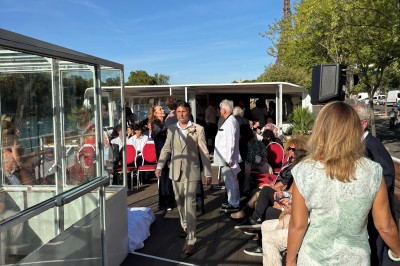 Intérieur d'un bateau avec éclairage vert, tables et chaises, vue sur une terrasse extérieure et des passants.