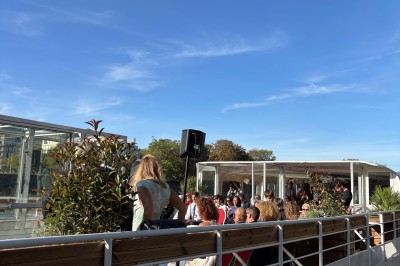 Intérieur d'un bateau avec éclairage vert, tables et chaises, vue sur une terrasse extérieure et des passants.