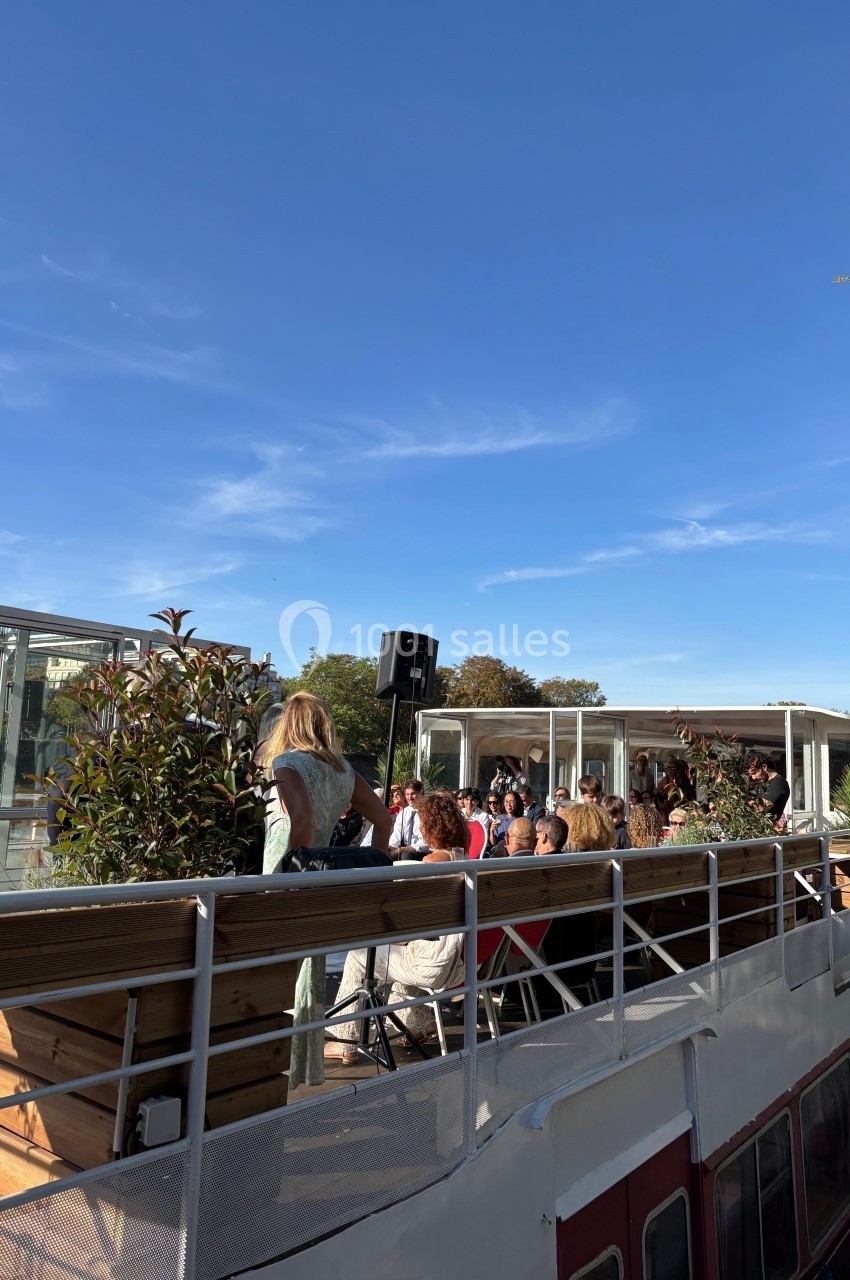 Des personnes assises sur le pont d'une péniche, entourées de plantes, sous un ciel bleu dégagé.