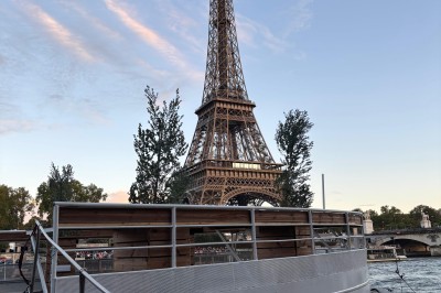 Intérieur d'un bateau avec éclairage vert, tables et chaises, vue sur une terrasse extérieure et des passants.
