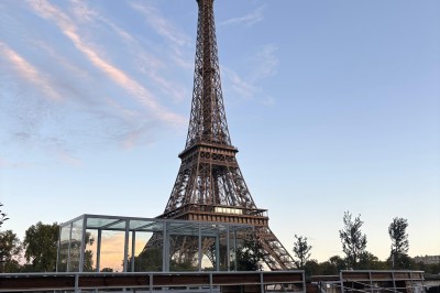 Intérieur d'un bateau avec éclairage vert, tables et chaises, vue sur une terrasse extérieure et des passants.