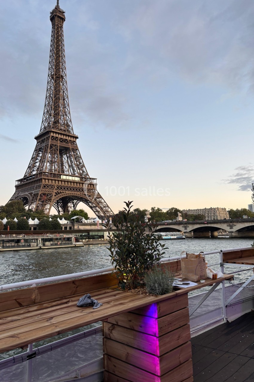 Vue de la tour Eiffel depuis une terrasse en bois au bord de la Seine, avec des plantes et un ciel légèrement nuageux.