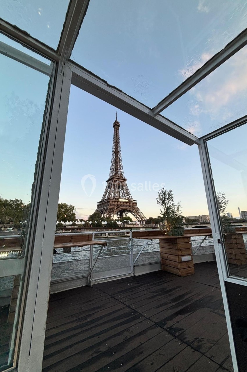 Vue de la tour Eiffel depuis une terrasse vitrée au bord de la Seine, entourée de végétation et d'un ciel dégagé.