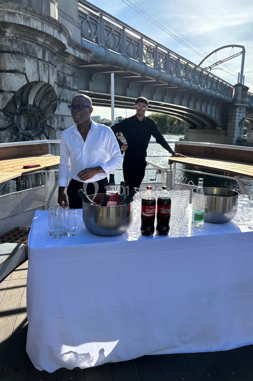 Deux hommes près d'une table dressée avec boissons, sur un bateau sous un pont par une journée ensoleillée.