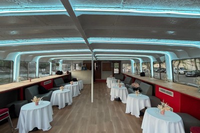 Intérieur d'un bateau avec éclairage vert, tables et chaises, vue sur une terrasse extérieure et des passants.