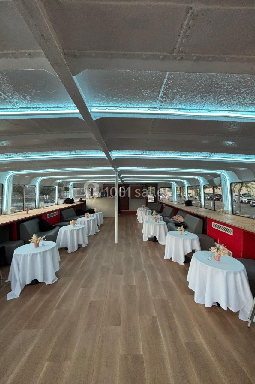 Intérieur d'un bateau aménagé avec des tables rondes décorées de fleurs et des banquettes le long des fenêtres.
