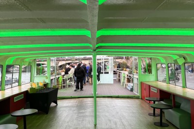 Intérieur d'un bateau avec éclairage vert, tables et chaises, vue sur une terrasse extérieure et des passants.