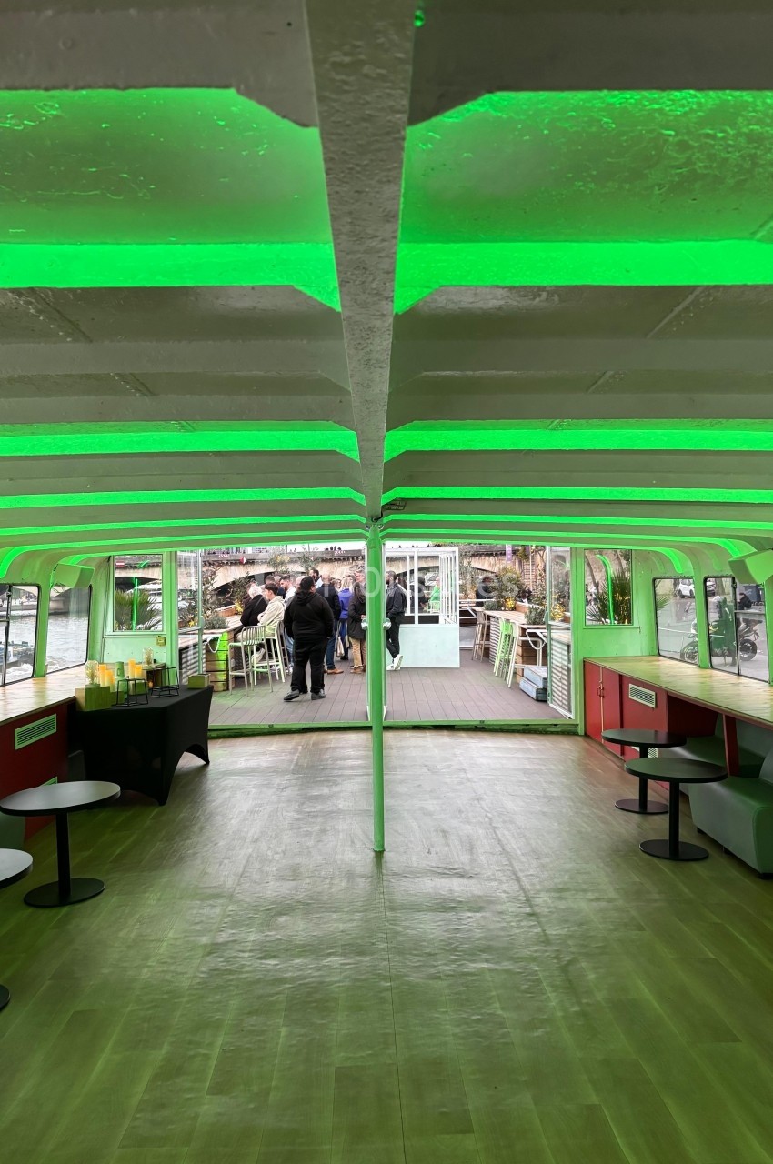 Intérieur d'un bateau avec éclairage vert, tables et chaises, vue sur une terrasse extérieure et des passants.