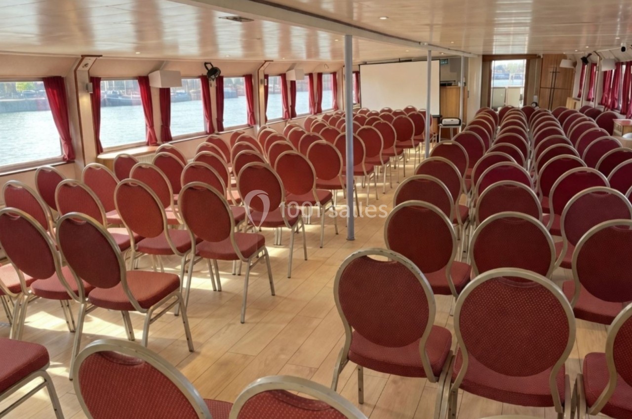 Salle de conférence sur un bateau avec des rangées de chaises rouges alignées et des fenêtres donnant sur l'eau.