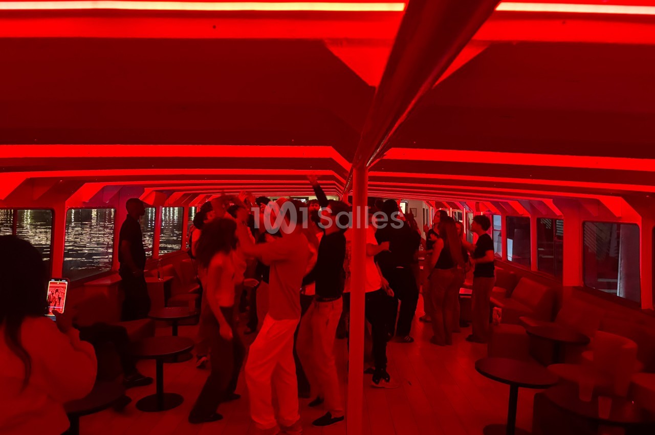 Des personnes dansent dans un espace intérieur éclairé par des lumières rouges, avec des tables et des banquettes autour.
