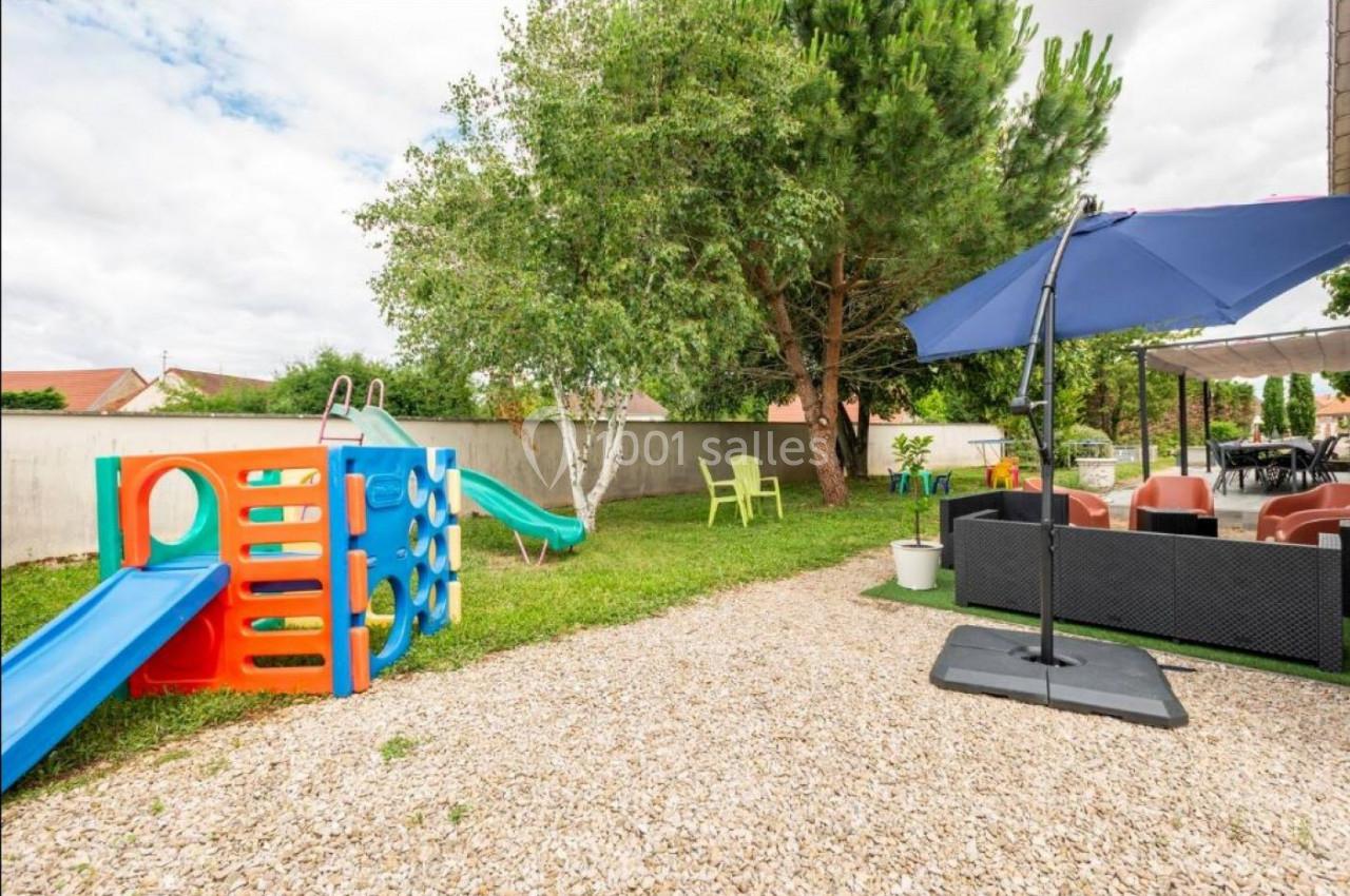 Aire de jeux extérieure avec toboggan coloré, pelouse, mobilier de jardin et parasol dans un espace clôturé.